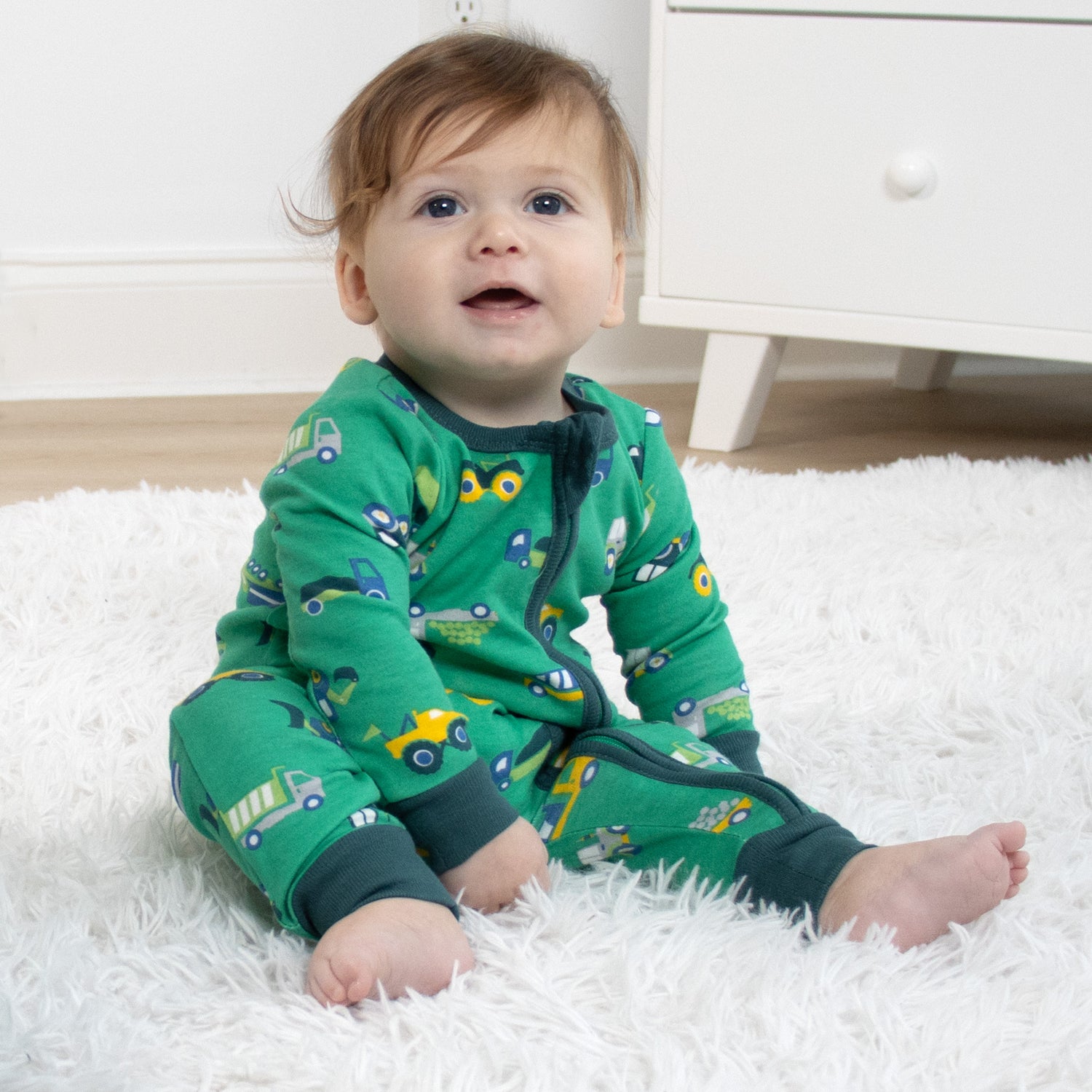 Baby wearing green pajamas with vehicle patterns sitting on a white rug.