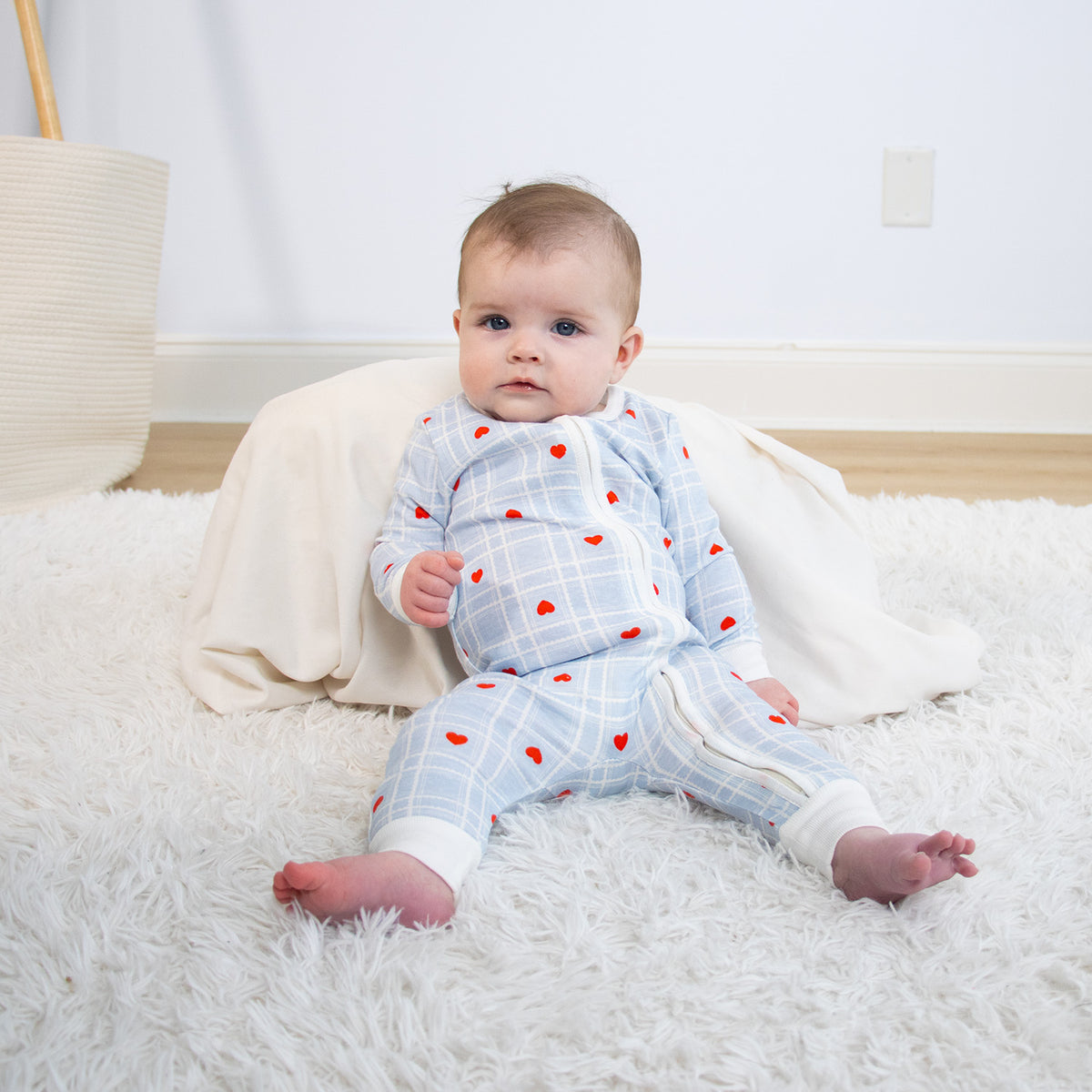Baby in a blue checkered outfit with red hearts sitting on a white fluffy rug.